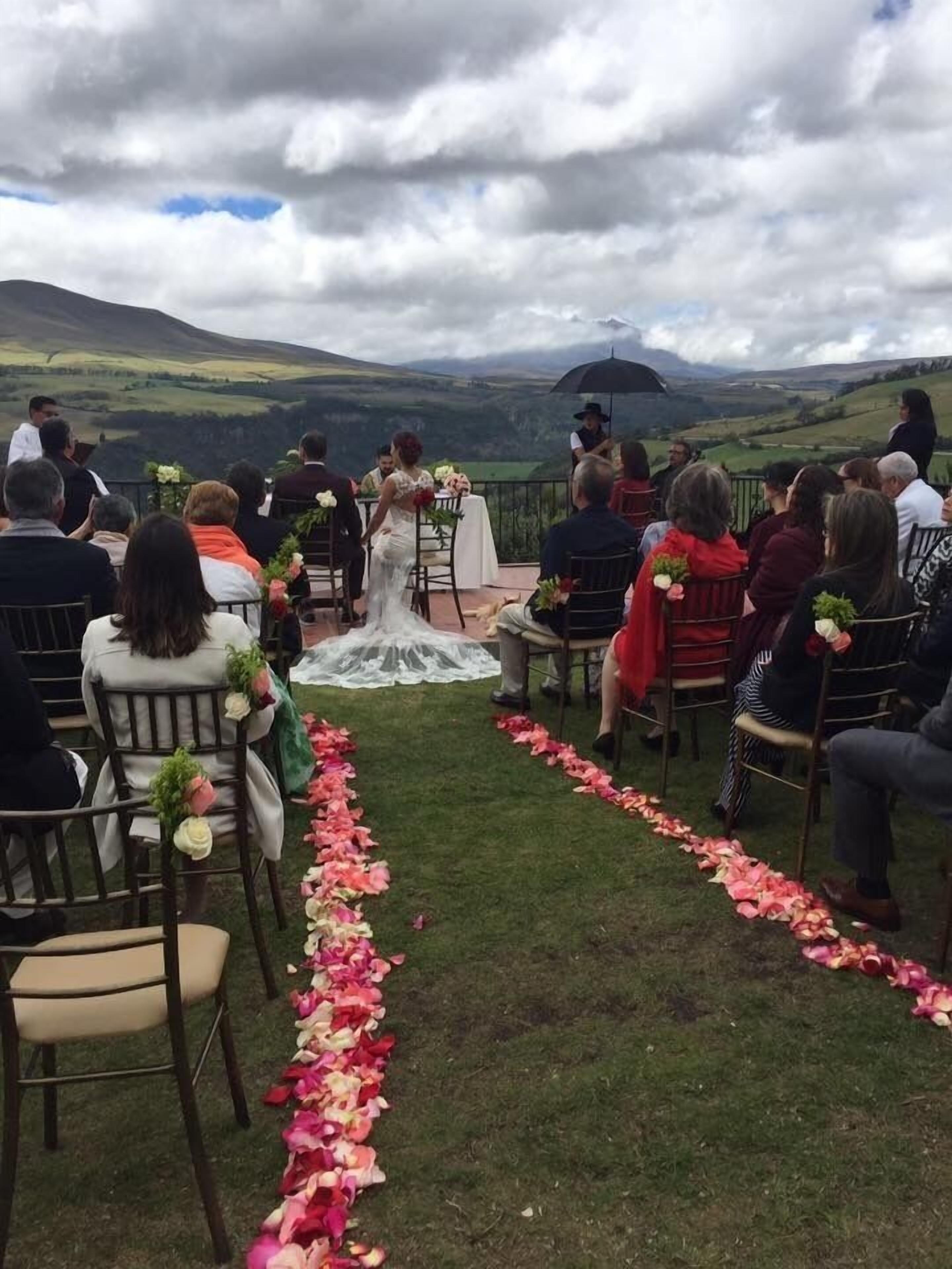 A wedding ceremony outdoors with guests seated facing the altar, a flower-decorated aisle, and a mountainous backdrop.