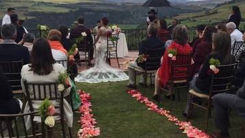 A wedding ceremony outdoors with guests seated facing the altar, a flower-decorated aisle, and a mountainous backdrop.