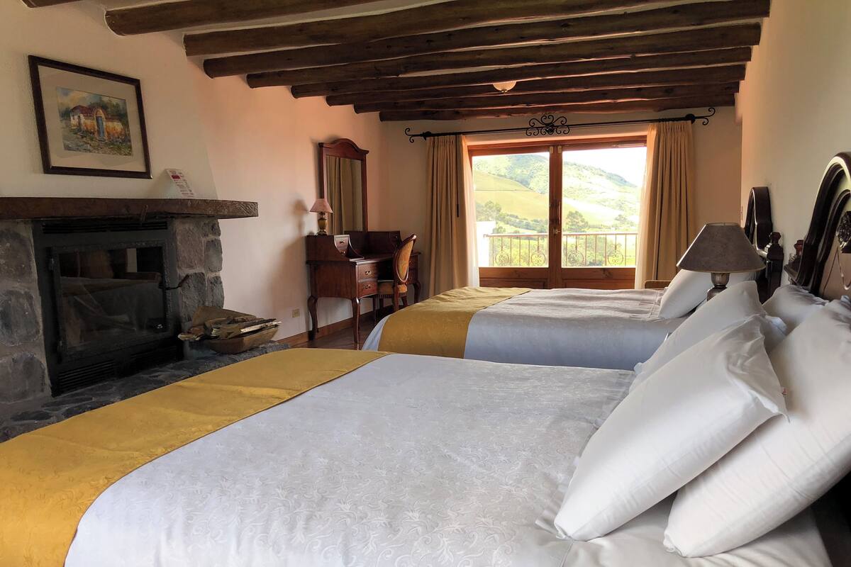 A room with two beds, a fireplace, a desk, and a view of the countryside.