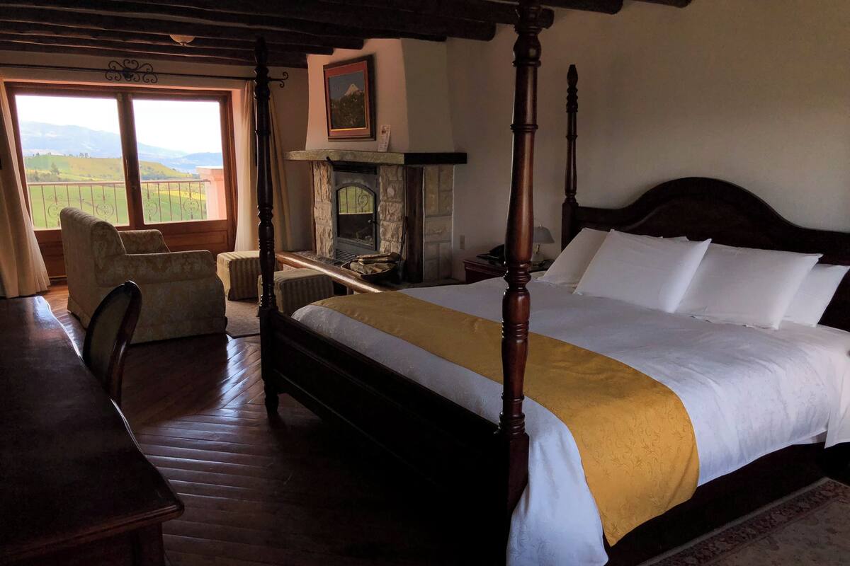 A four-poster bed with a stone fireplace, a view of the countryside through a large window, and a wooden desk.
