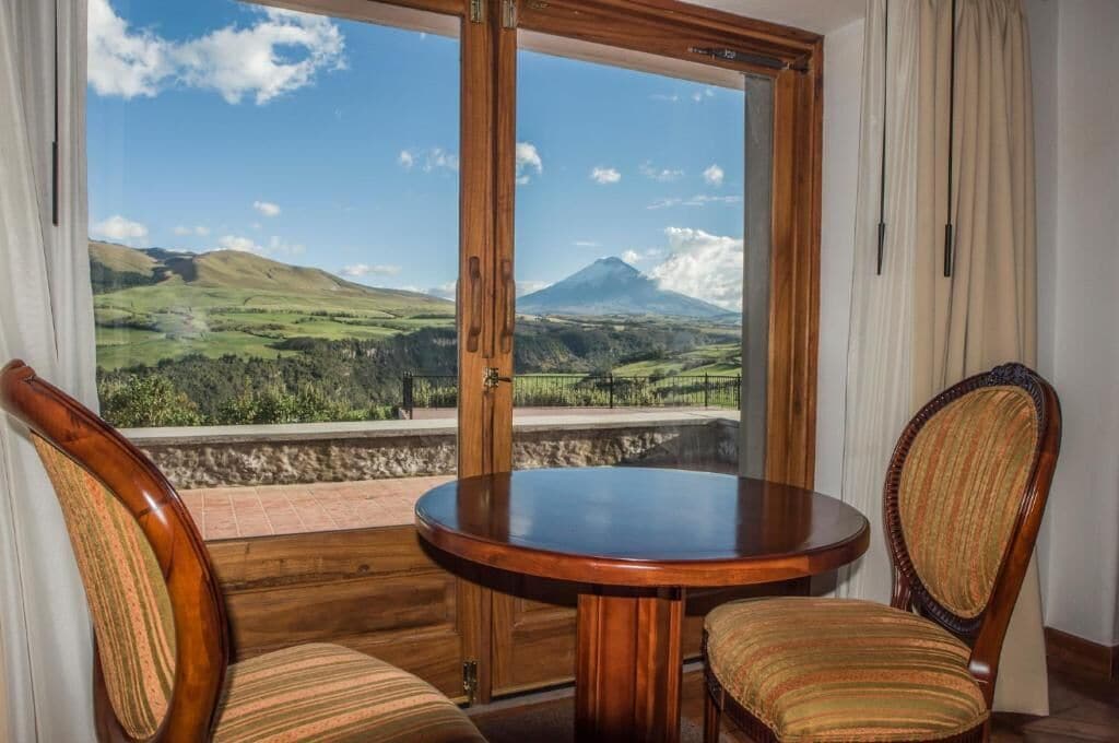 Una habitación con una mesa de madera y dos sillas tapizadas, un ventanal con vistas a una montaña y paisaje verde.