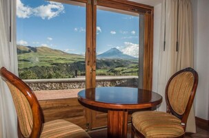 A room with a wooden table and two upholstered chairs, a large window with a view of a mountain and green landscape.