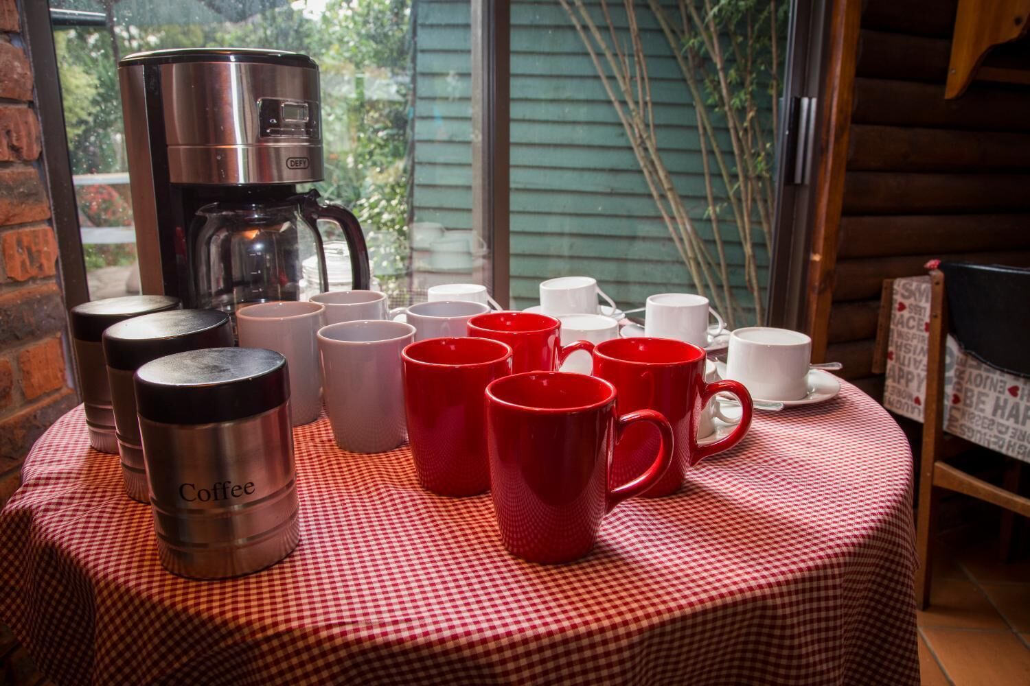 Een tafel met een koffiezetapparaat en verschillende mokken, een geruit tafelkleed en uitzicht op groen door een raam.