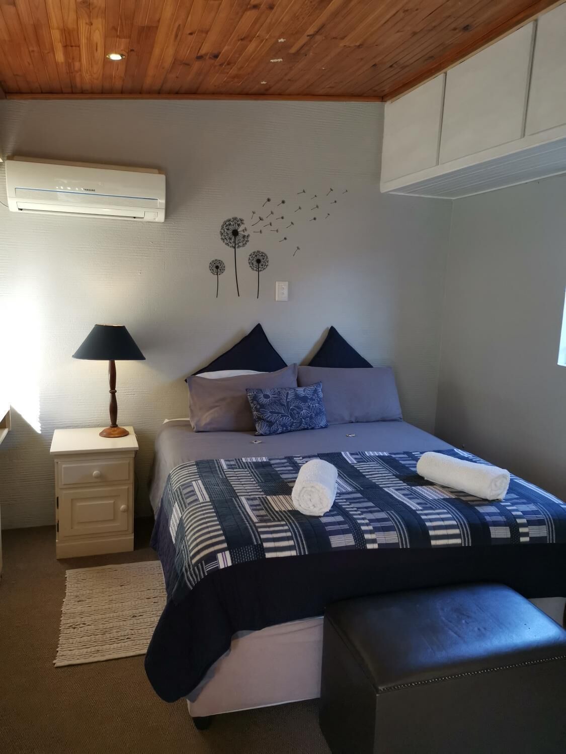 A bedroom with a bed, bedside table, lamp, air conditioning unit, and wall art.