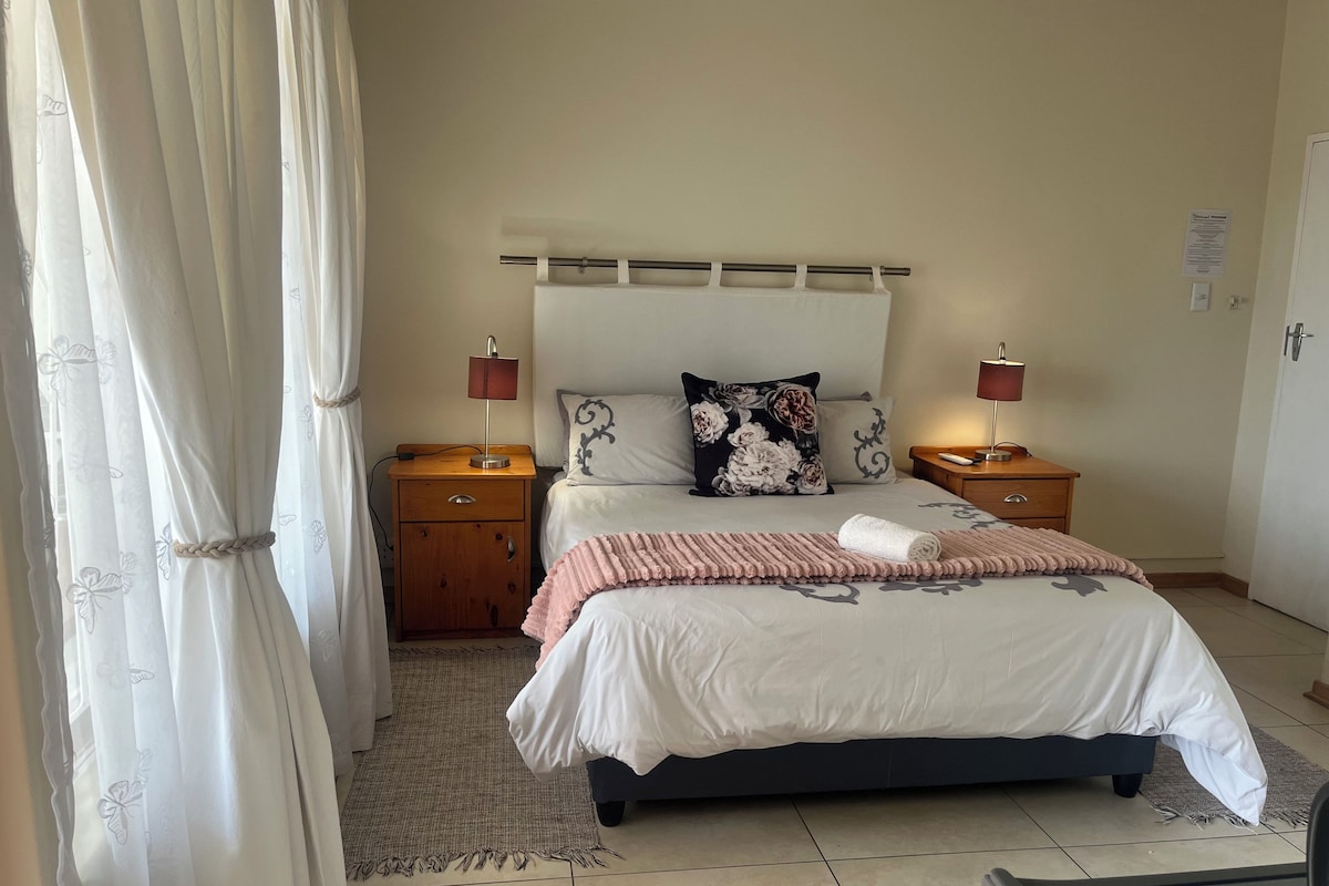 Ein Schlafzimmer mit einem Bett, zwei Nachttischen, einer Lampe, einem Kissen, einer Decke und einem Vorhang.