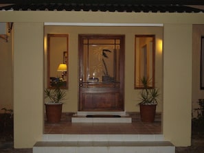 Une porte en bois avec un panneau de verre et un motif représentant un palmier, flanquée de deux plantes en pot et d’une petite marche menant à la porte.