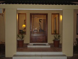 Uma porta de madeira com um painel de vidro e um desenho de palmeira, adornada por duas plantas em vasos e um pequeno degrau que leva à porta.