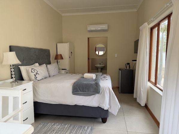 Un dormitorio con cama, mesitas de noche, una lámpara, un espejo, un lavamanos y una ventana con cortinas.