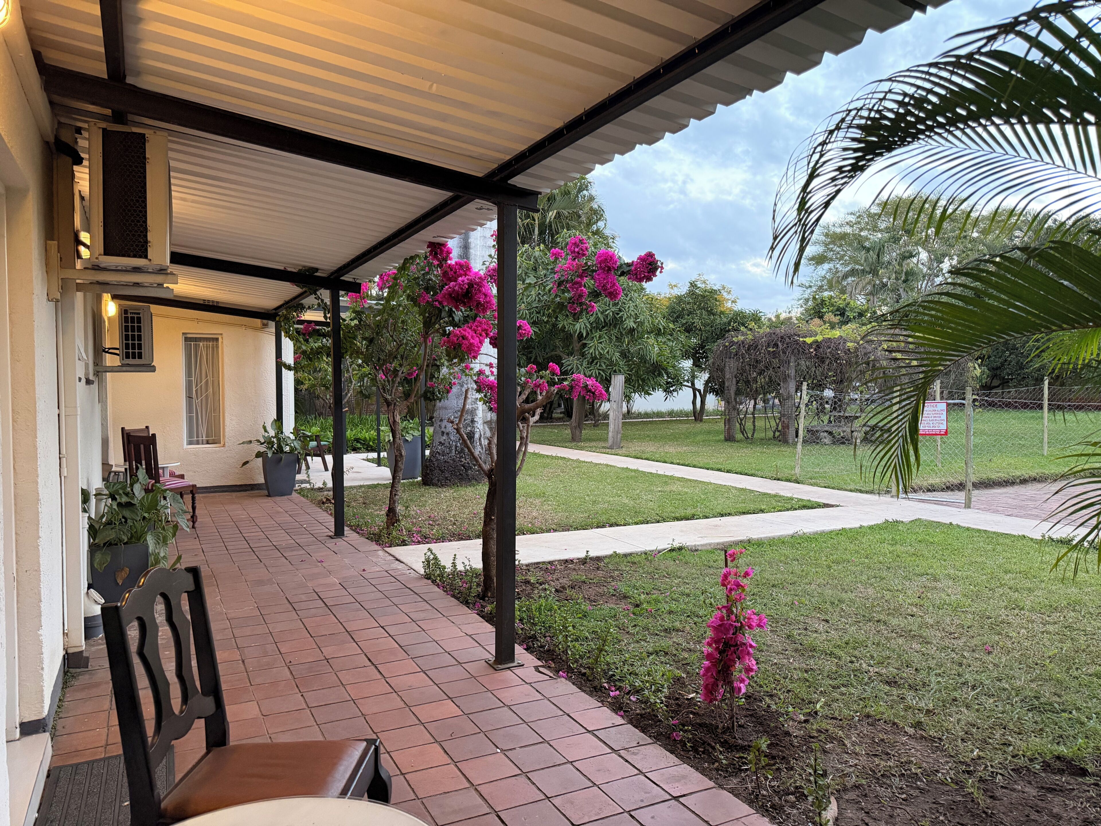 A covered outdoor area with a patio, chairs, and a view of a grassy area with trees and a fence.