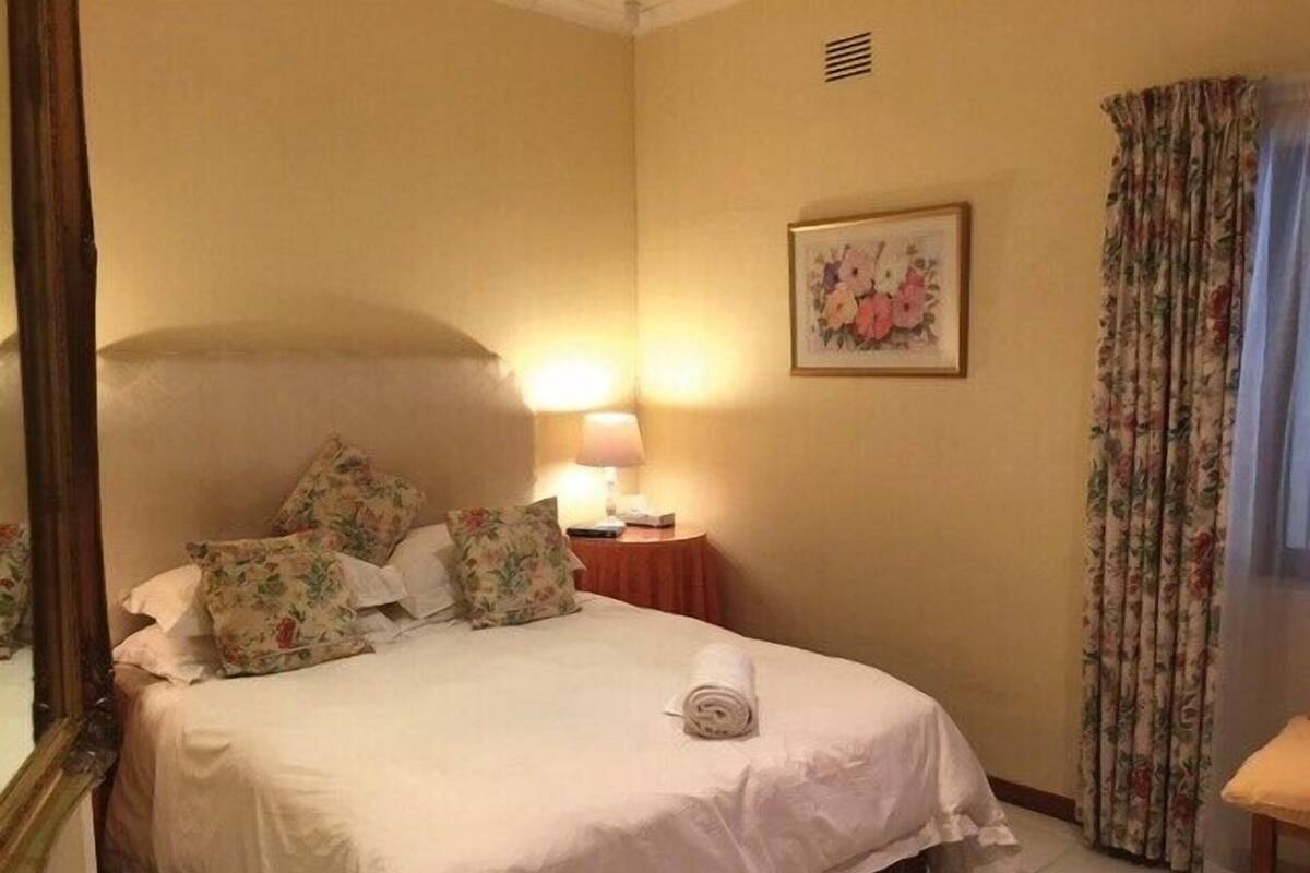 Een netjes opgemaakt bed met bloemenkussens, een bedlampje en een lijst met bloemwerk aan de muur.