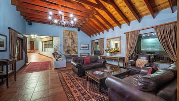 A spacious living room with a high wooden ceiling, leather sofas, a coffee table, and a large window with curtains.