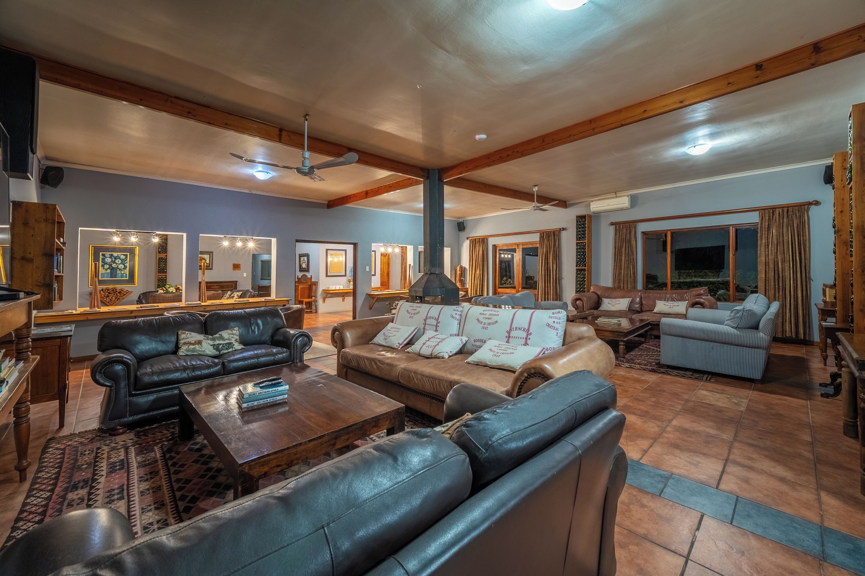 A spacious living room with leather sofas, a coffee table, and a fireplace. There are ceiling fans and a large window with curtains.