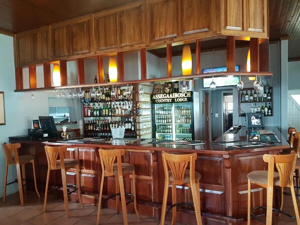 Zona de bar con mostradores de madera, varios taburetes y una estantería repleta de licores.