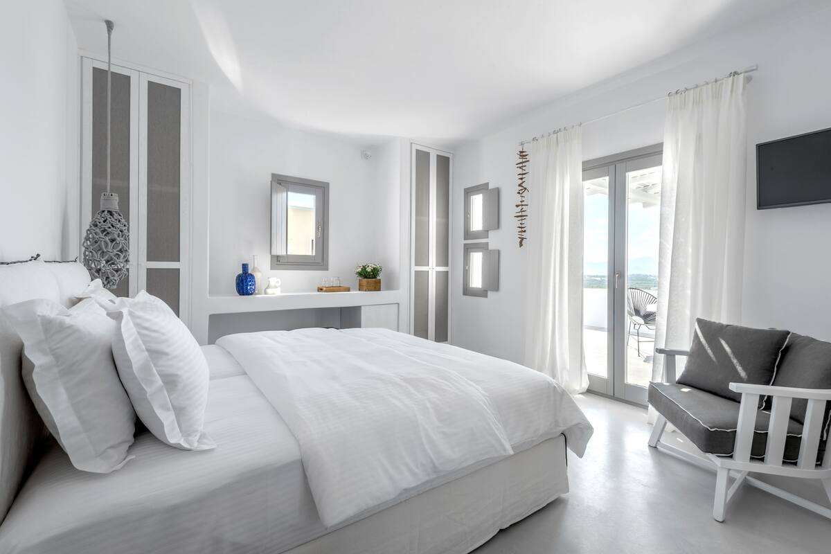 A modern bedroom with a bed, a white chair, a small table with a mirror, and a view through a glass door.