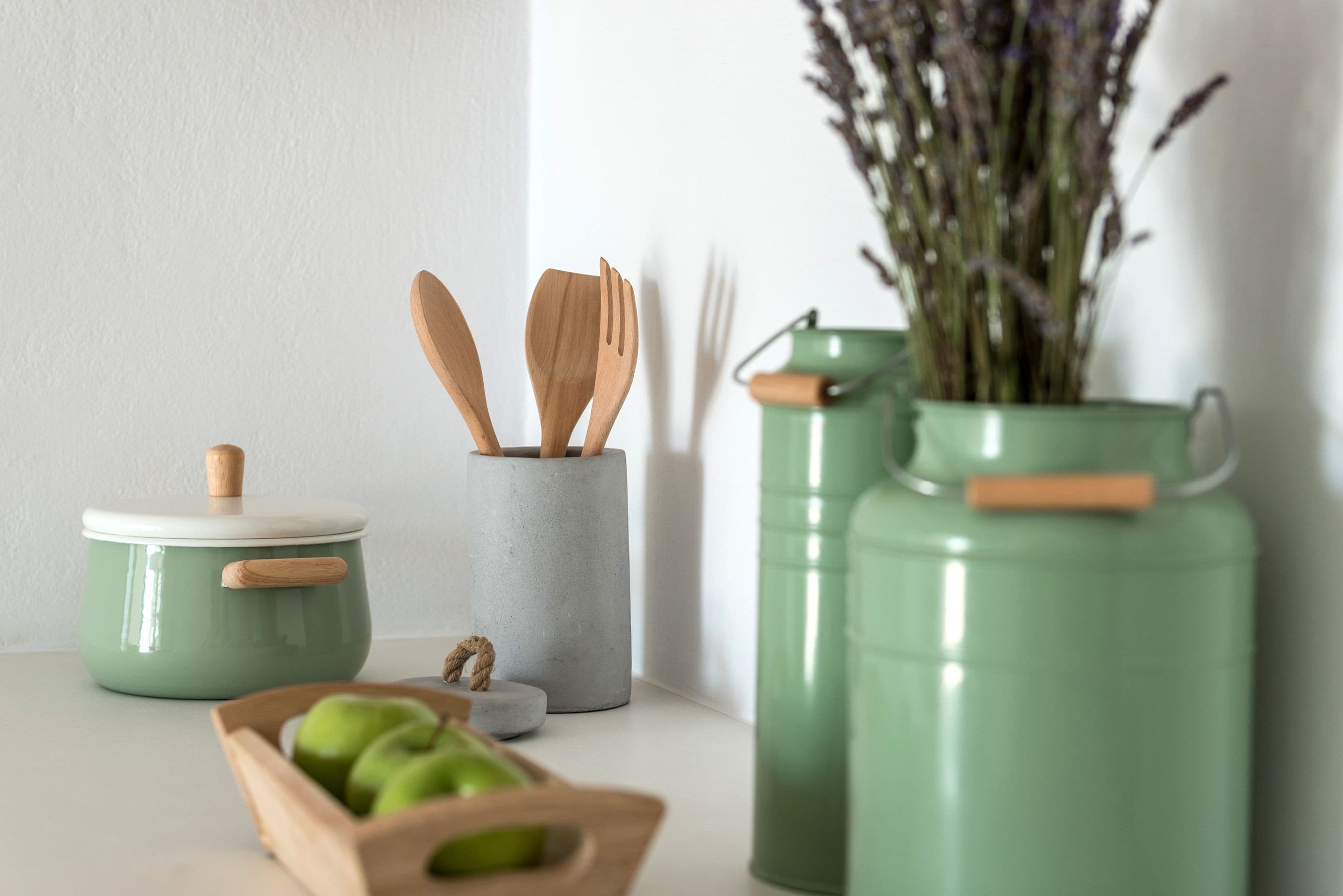 A kitchen area with a green pot, wooden utensils, a bowl of green apples, and a vase with lavender.