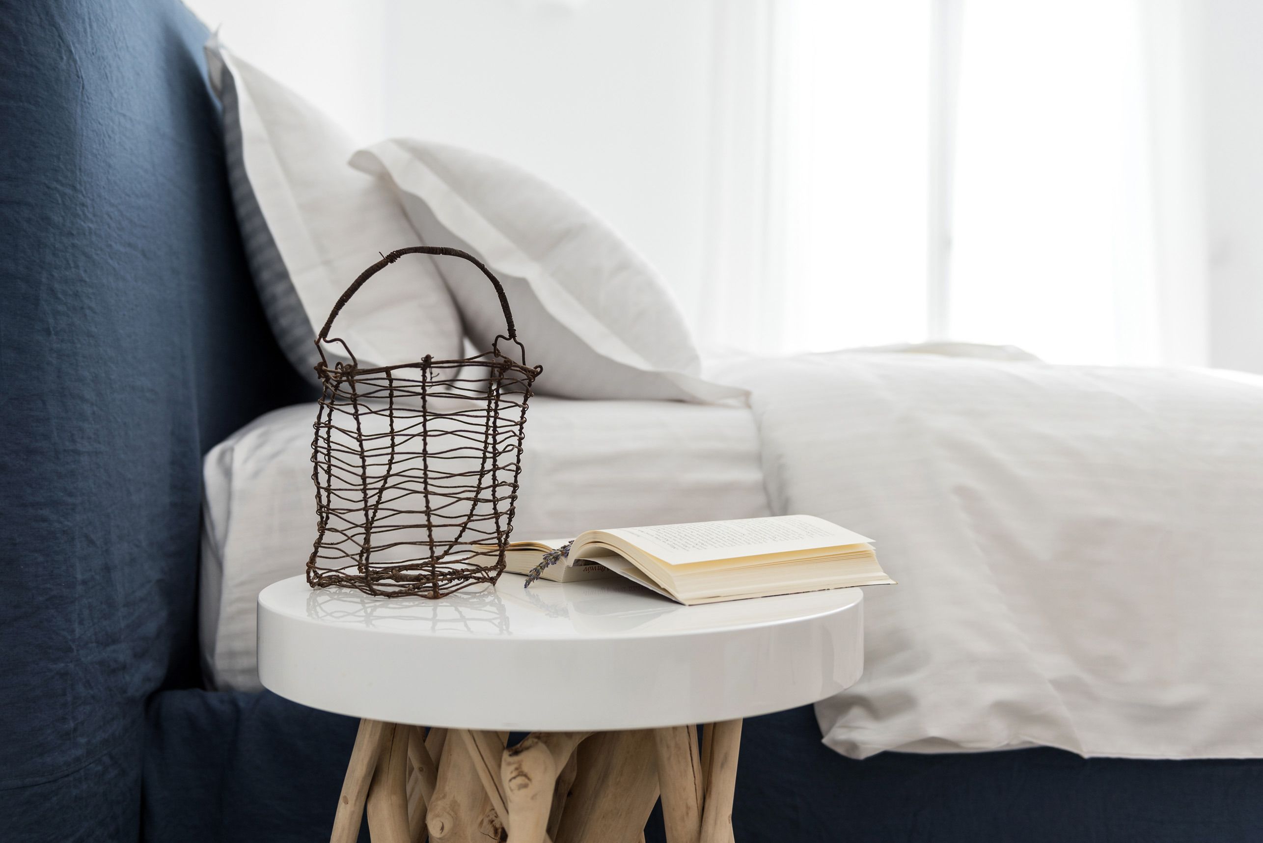 A round white table with a wire basket and an open book on it, placed on a blue couch with white pillows.