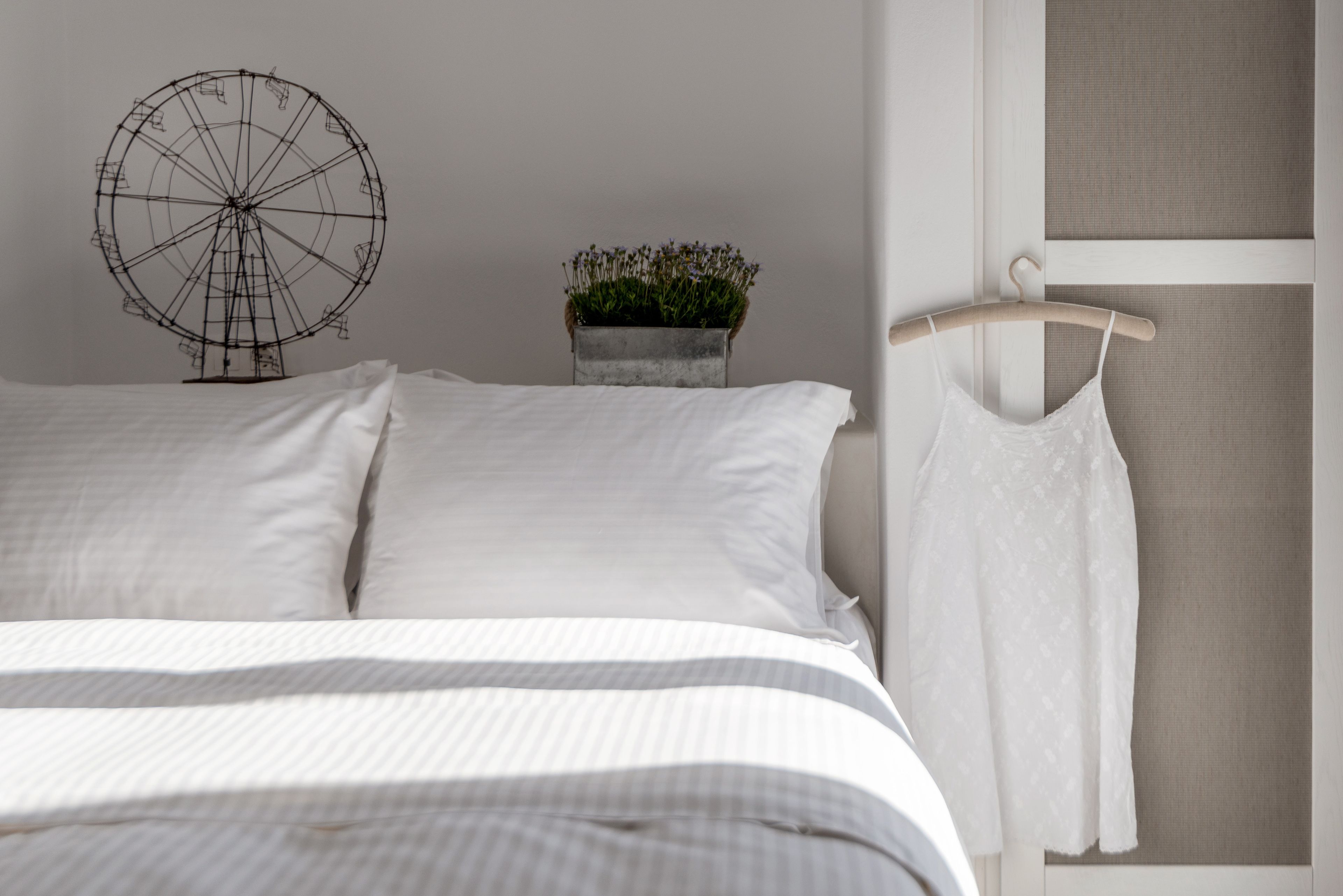 A neatly made bed with white linens, a small potted plant, and a white dress on a hanger.
