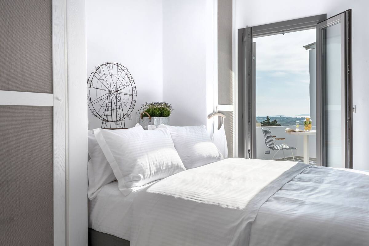 A modern bedroom with a bed, a decorative wall piece, and a view of a balcony with outdoor furniture.
