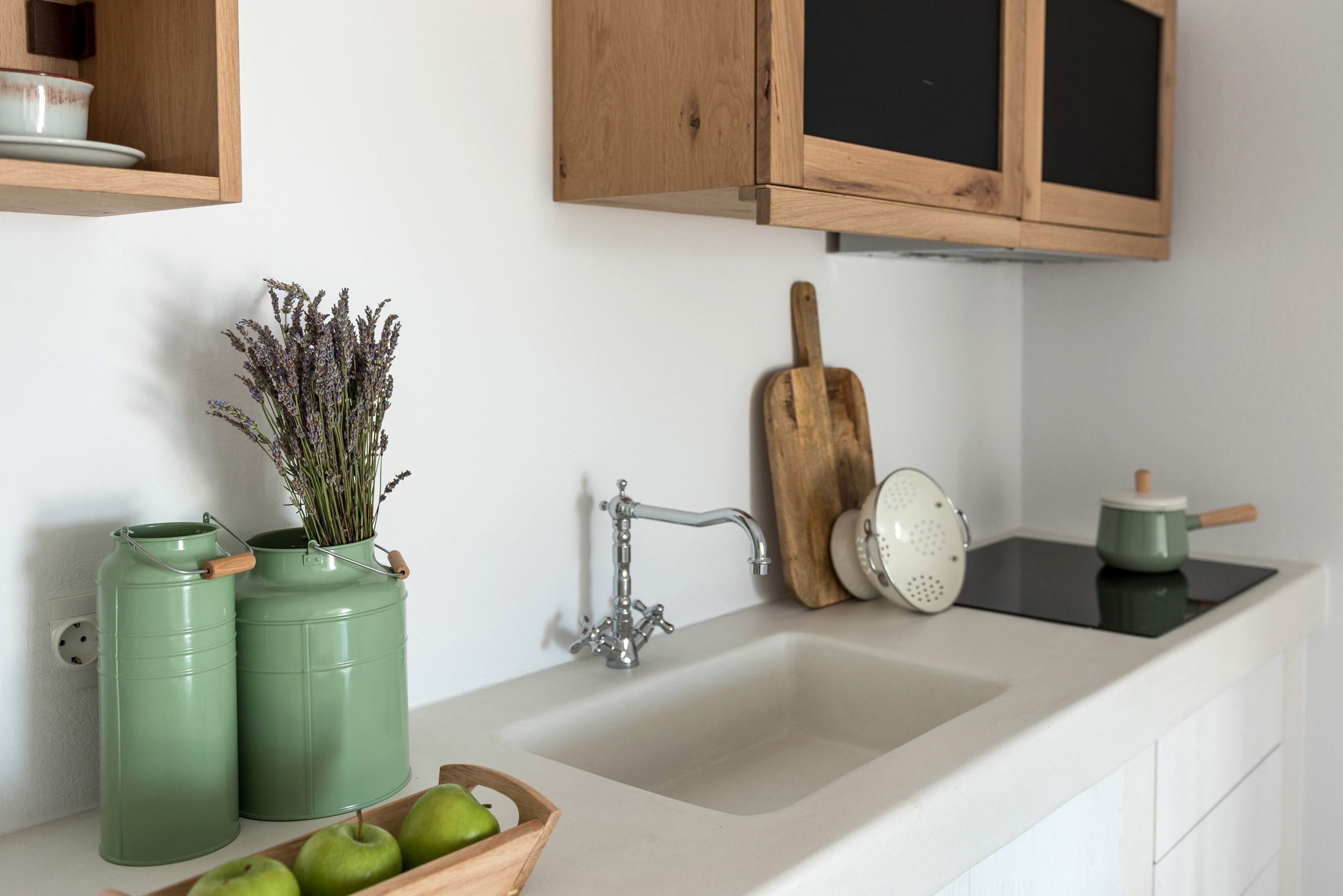 A kitchen with a white sink, wooden cabinets, a green pot, and a bowl of green apples.