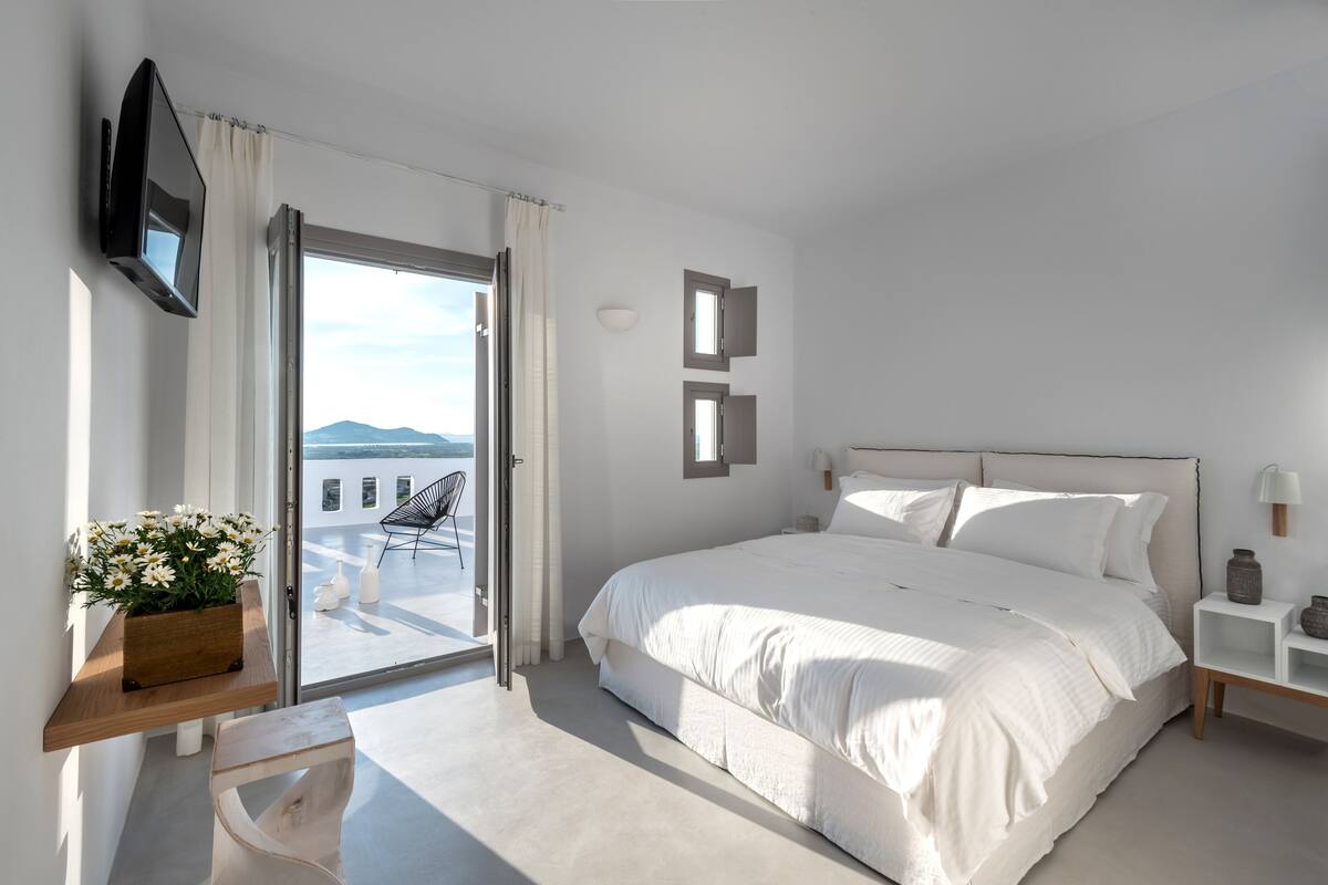 A modern bedroom with a large bed, a TV mounted on the wall, a balcony with a chair, and a view of the mountains.