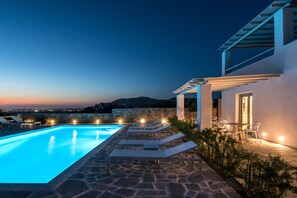 A swimming pool with lounge chairs and a table, surrounded by stone walls and a view of the city at dusk. - Seven Suites (Naxos)