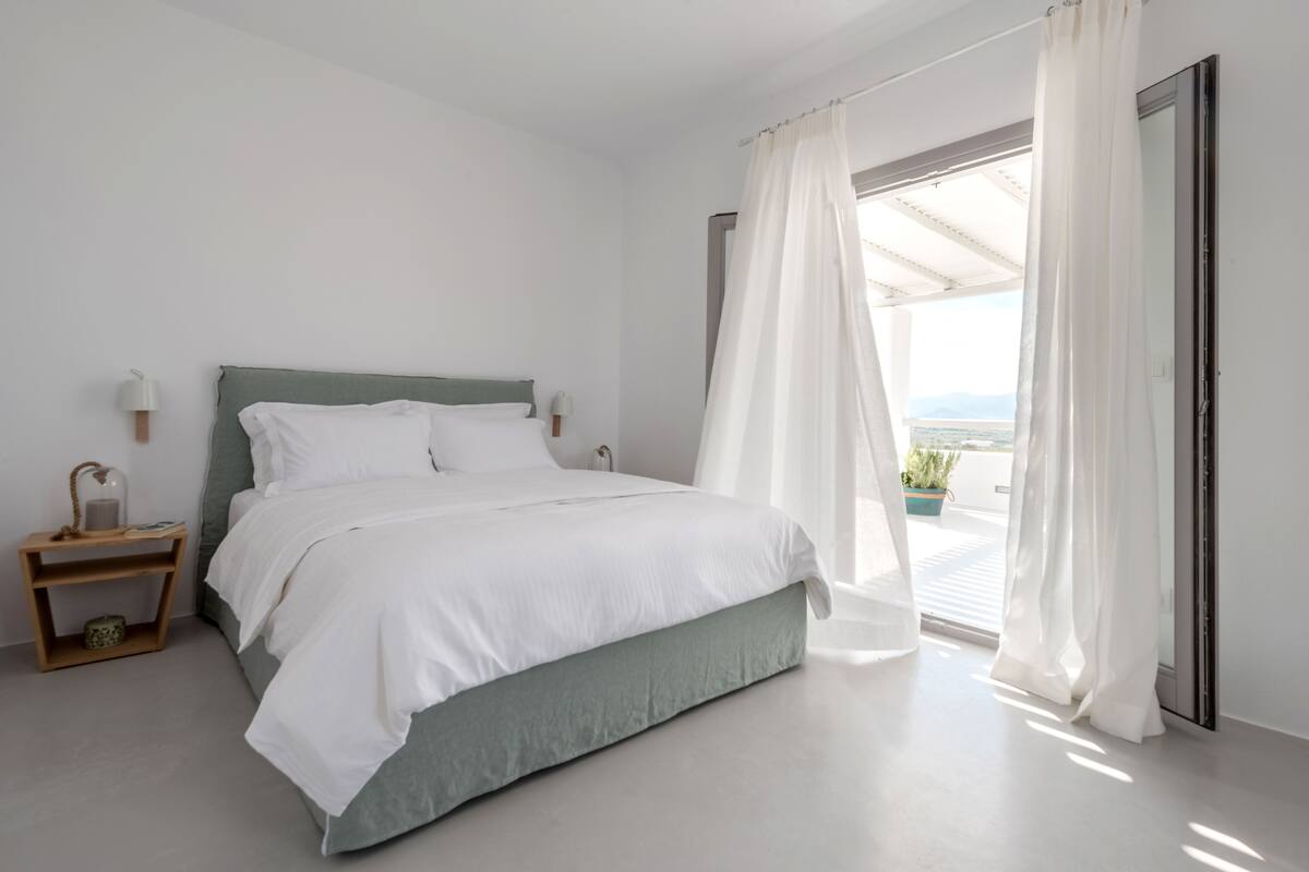 A modern bedroom with a bed, a nightstand, a window with sheer curtains, and a view of the outdoors.