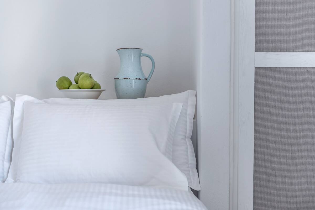A neatly made bed with white bedding, a bowl of green apples, and a light blue pitcher on a bedside table.