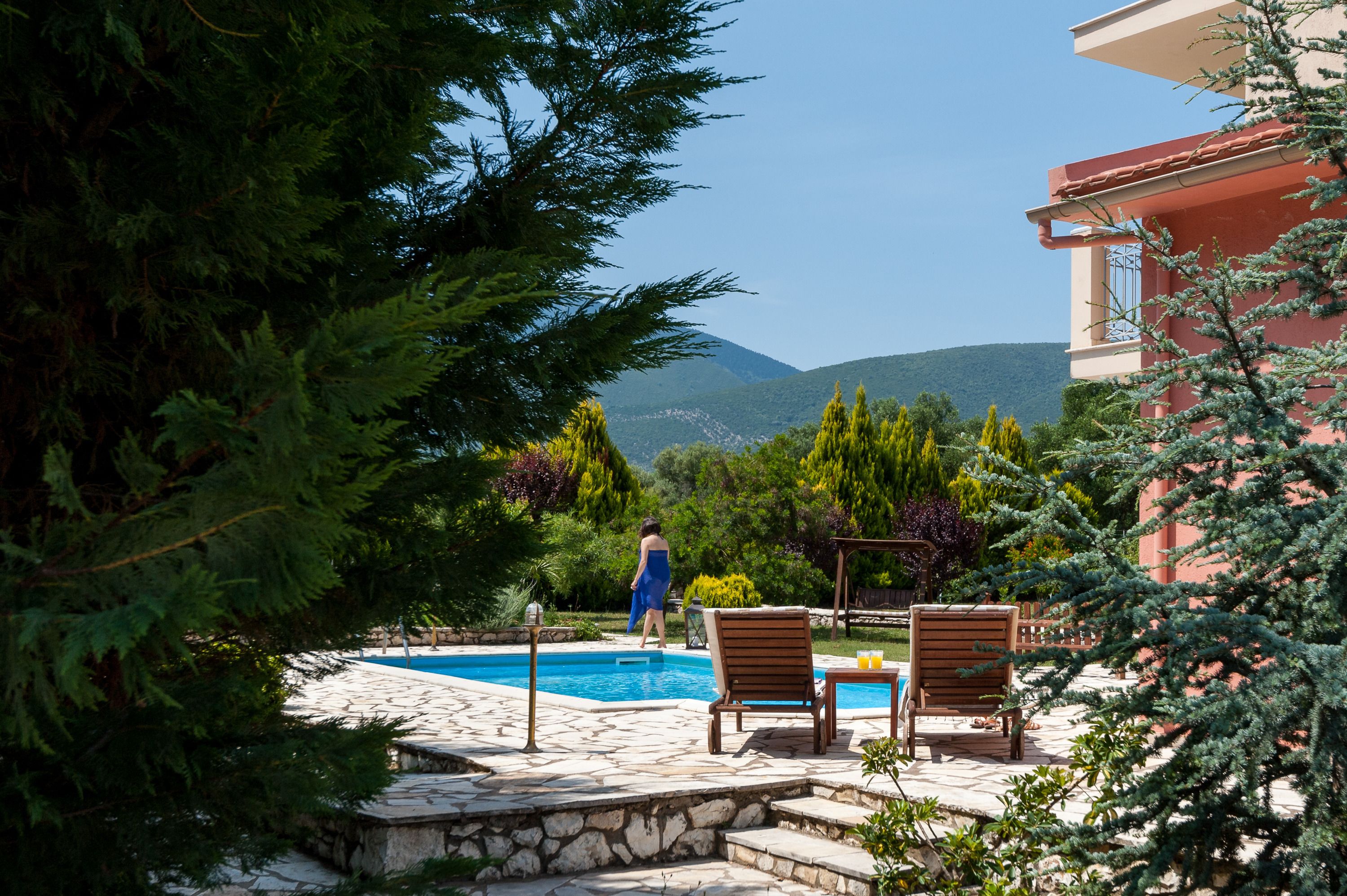 A pool area with stone tiles, wooden lounge chairs, and a person in a blue dress.