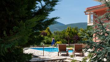 A pool area with stone tiles, wooden lounge chairs, and a person in a blue dress.