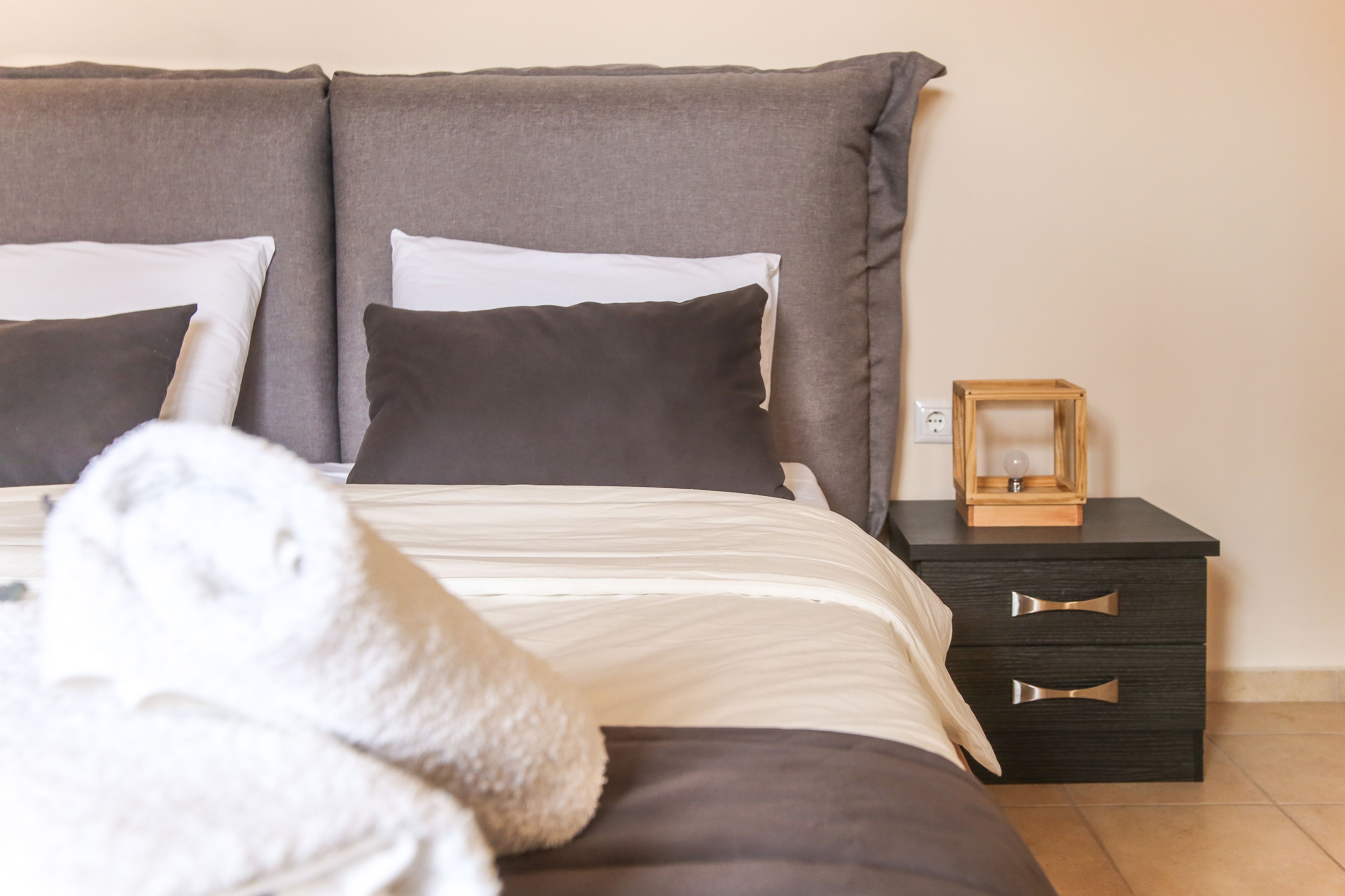 Una cama bien hecha con una colcha blanca y dos almohadas, una mesita de noche pequeña con una lámpara, y una cómoda de madera con cajones.