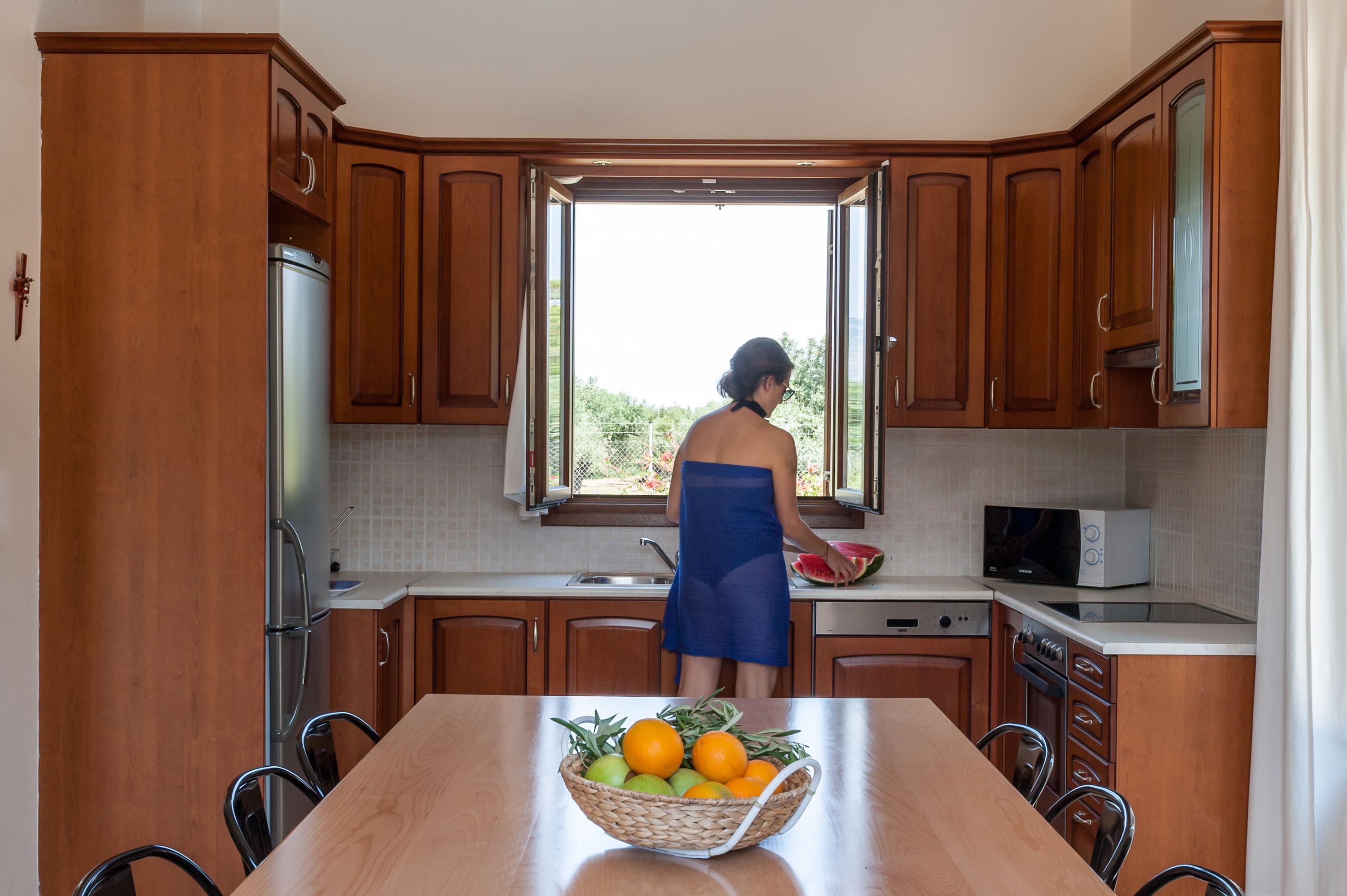 Une cuisine avec des meubles en bois, un réfrigérateur en acier inoxydable, un four à micro-ondes et une table à manger agrémentée d’un panier de fruits.