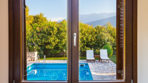 A sliding glass door with a view of a swimming pool, two lounge chairs, and a stone patio. - Penelope Villas (Kefalonia)