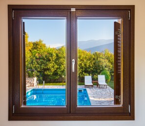 A sliding glass door with a view of a swimming pool, two lounge chairs, and a stone patio. - Penelope Villas (Kefalonia)