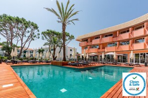 Ein Poolbereich mit Liegestühlen und Sonnenschirmen, umgeben von Palmen und einem Gebäude mit Balkonen.