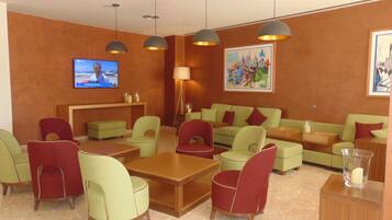 Una hall d'albergo con televisore, divani verdi, poltrone rosse, un tavolino in legno, lampade a sospensione e un quadro appeso al muro.