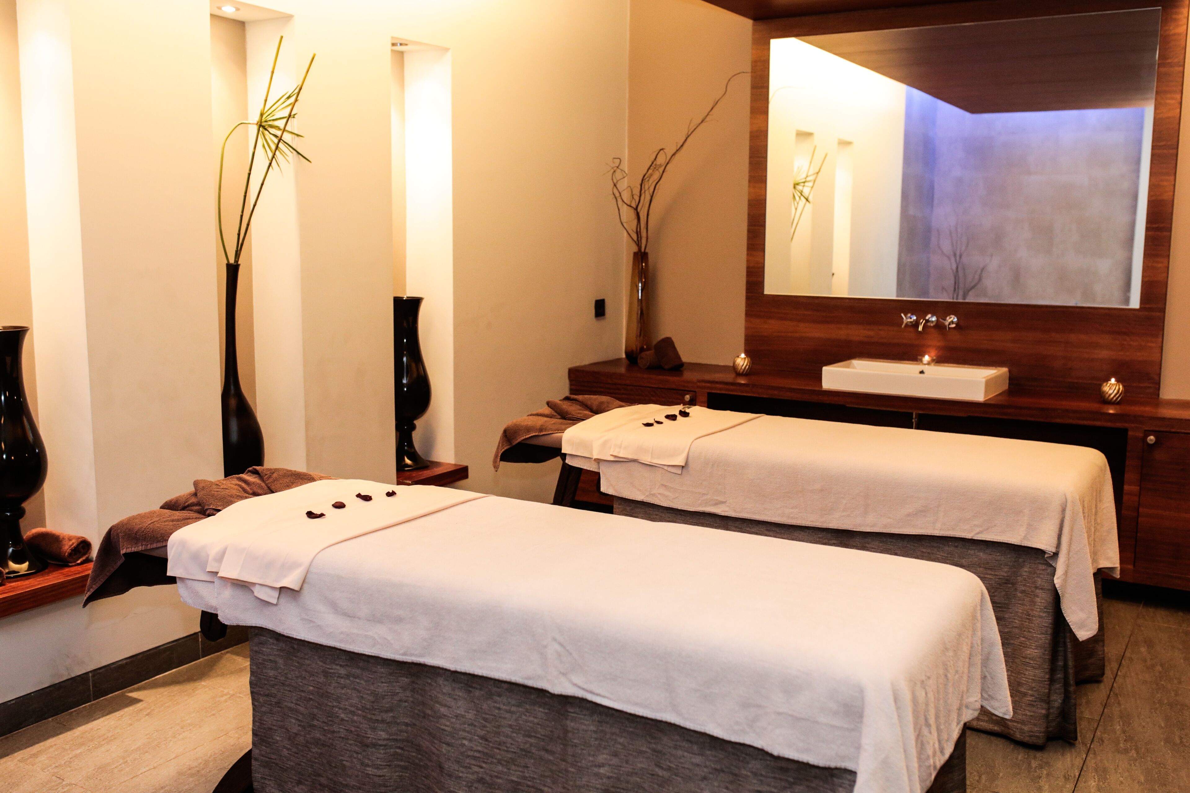 A spa room with two massage tables, a wooden vanity with a mirror, and two vases with branches.