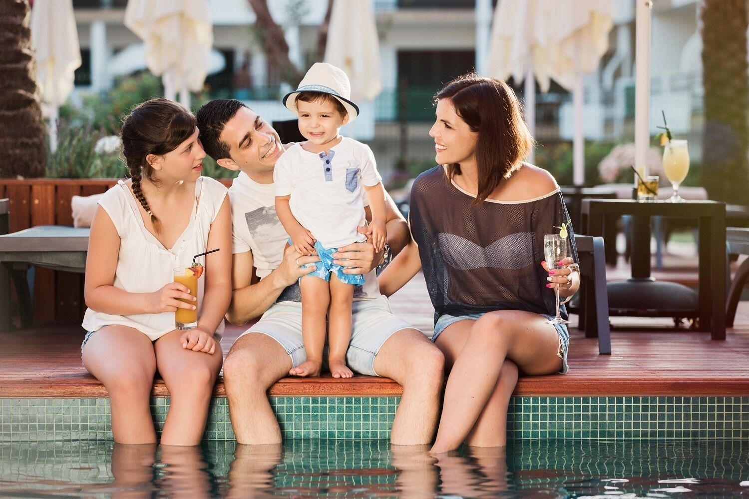 Uma família de quatro pessoas sentada à beira da piscina, com bebidas, em um ambiente com mobiliário de exterior e piscina revestida de azulejos.