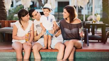 Uma família de quatro pessoas sentada à beira da piscina, com bebidas, em um ambiente com mobiliário de exterior e piscina revestida de azulejos.