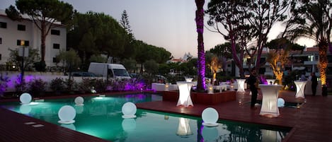 A pool area with a wooden deck, a palm tree, and tables with white lighting.