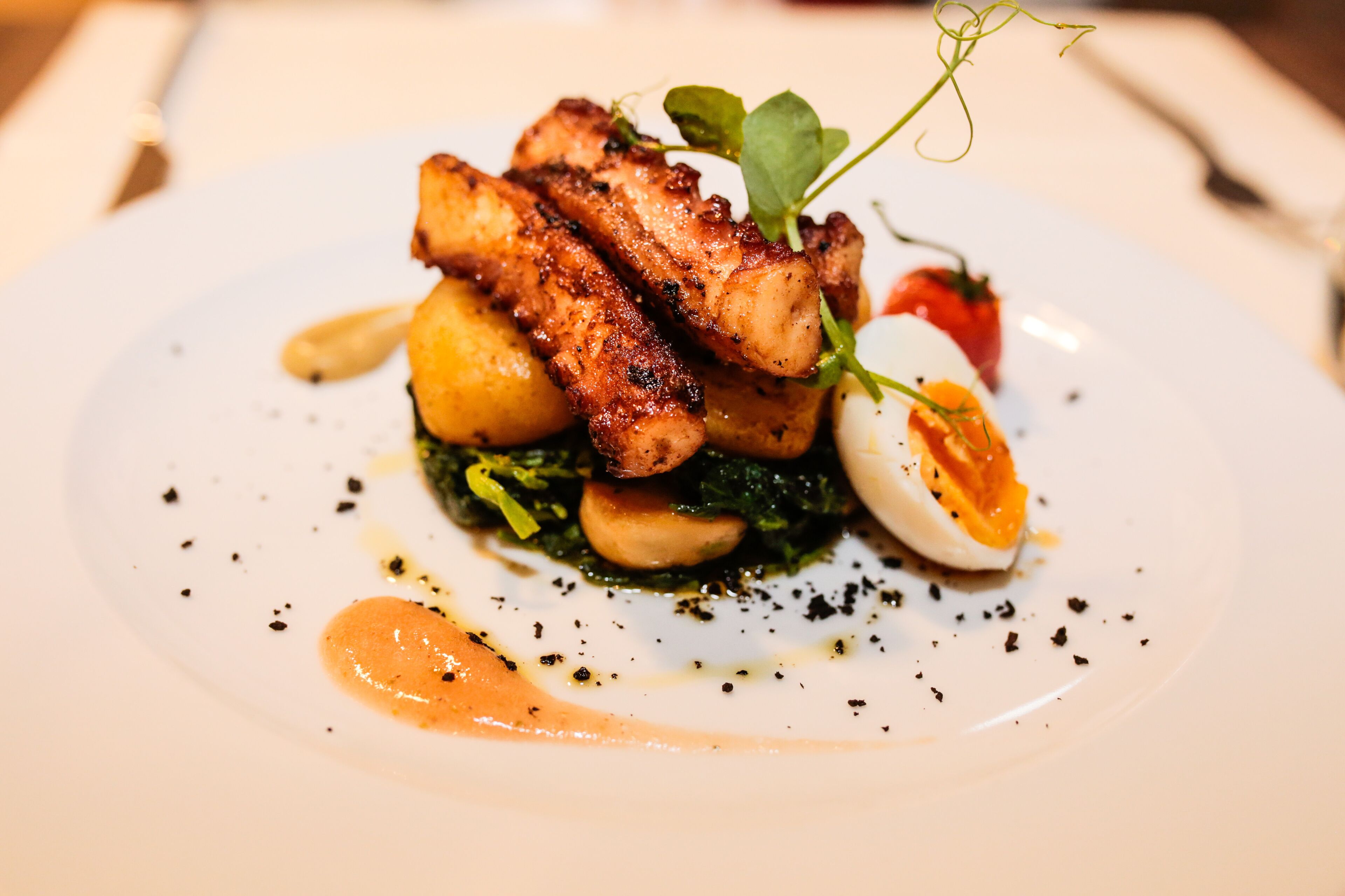 A plate of food with grilled meat, potatoes, greens, and a boiled egg, served on a white plate with a sauce and black pepper.