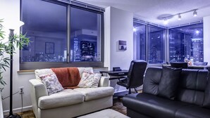 Una sala moderna con un ventanal que ofrece vista a la ciudad, un sofá blanco con cojines estampados, un sofá de cuero negro y un comedor con mesa y sillas.