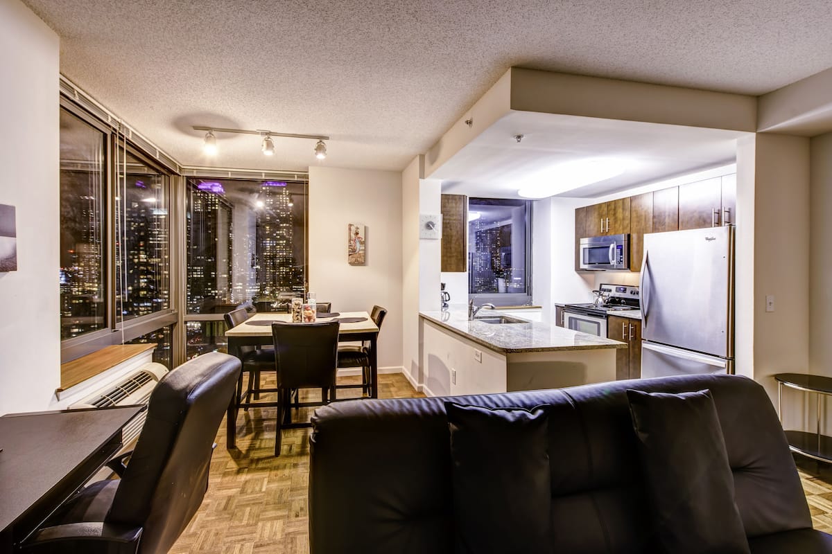 Una cocina moderna con microondas, estufa y refrigerador. Hay un comedor con mesa y sillas, y un amplio ventanal con vista a la ciudad.