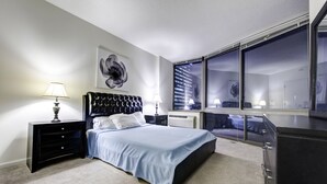 Un dormitorio moderno con una cama grande, una mesita de noche, un aparador y vistas a un paisaje urbano a través de un ventanal amplio.
