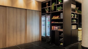 Moderni hotellihuone, jossa on puupanelilla verhoiltu seinä, musta hyllykkö, jossa on kasveja ja pientä purtavaa, sekä juomia sisältävä jääkaappi.