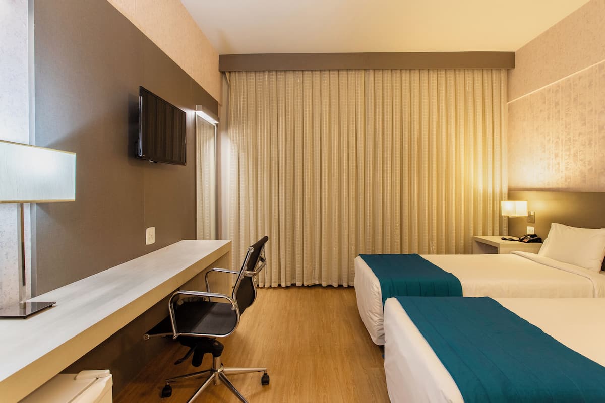 Een hotelkamer met een bureau, twee bedden, een televisie en een groot raam met gordijnen.