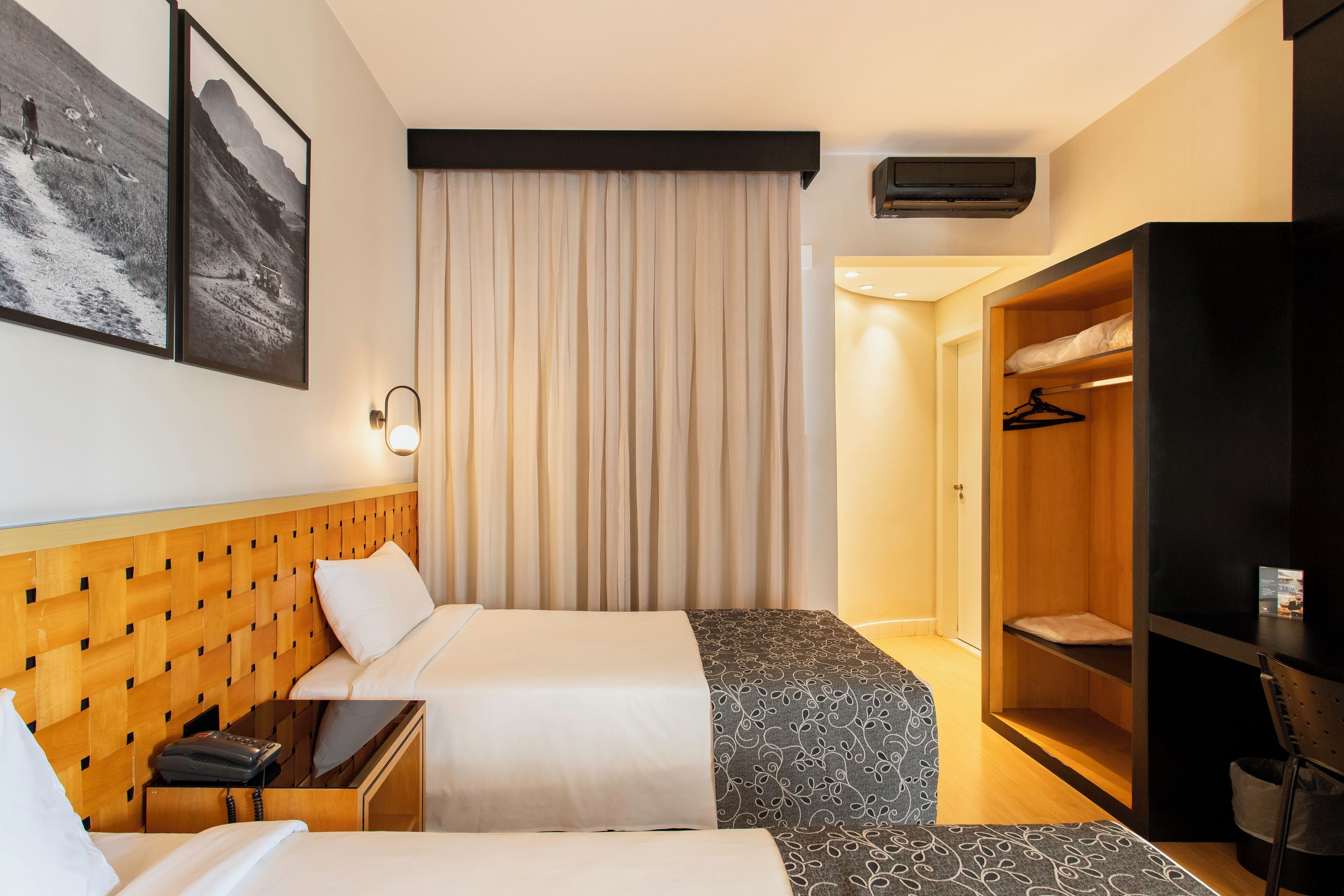 Quarto de hotel com duas camas, cabeceira de madeira, guarda-roupa e ar condicionado fixado na parede.