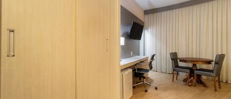 A modern room with a wooden desk, a small refrigerator, a TV mounted on the wall, and a dining table with chairs.