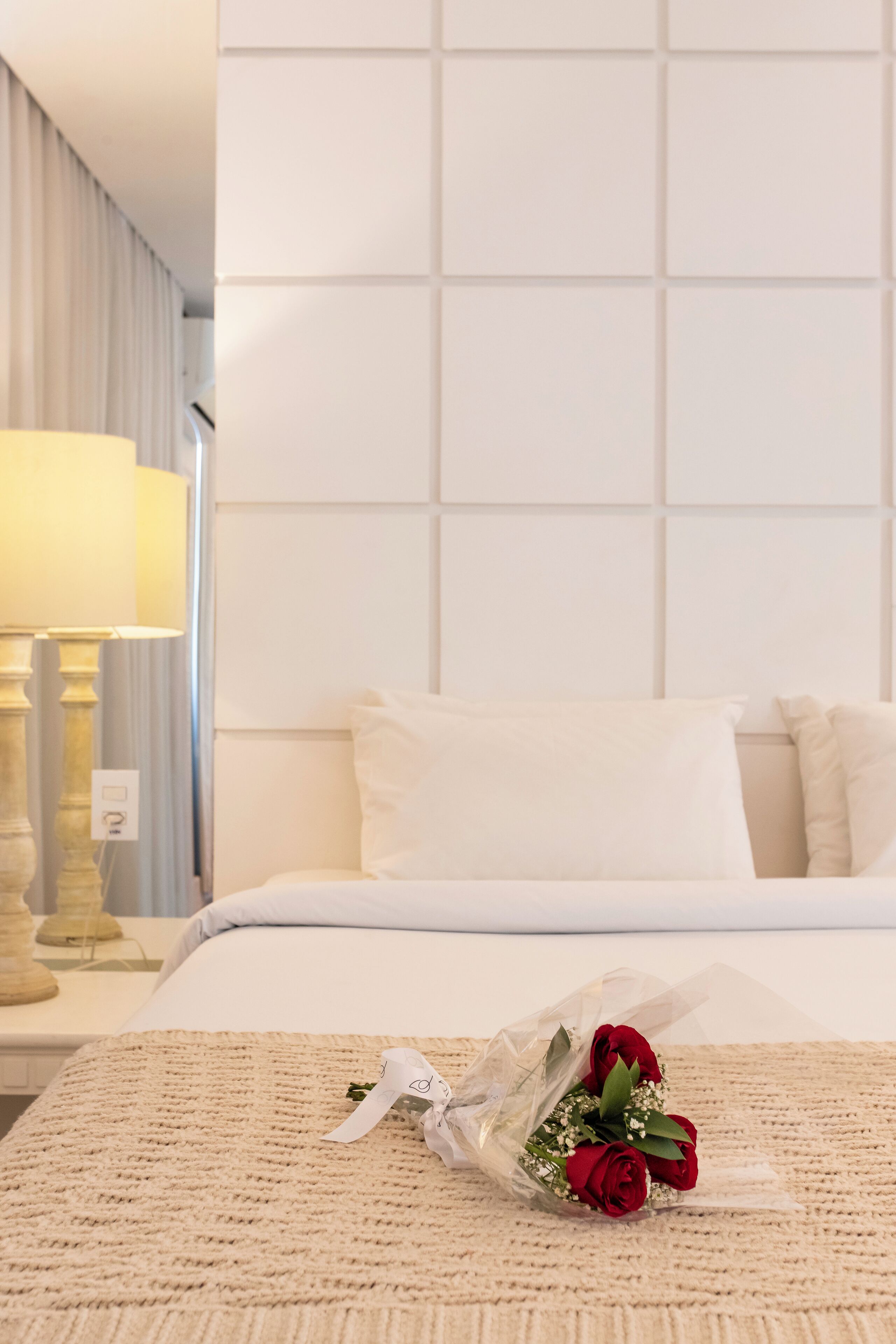 A hotel room with a neatly made bed, a bedside lamp, and a bouquet of red roses on the bed.