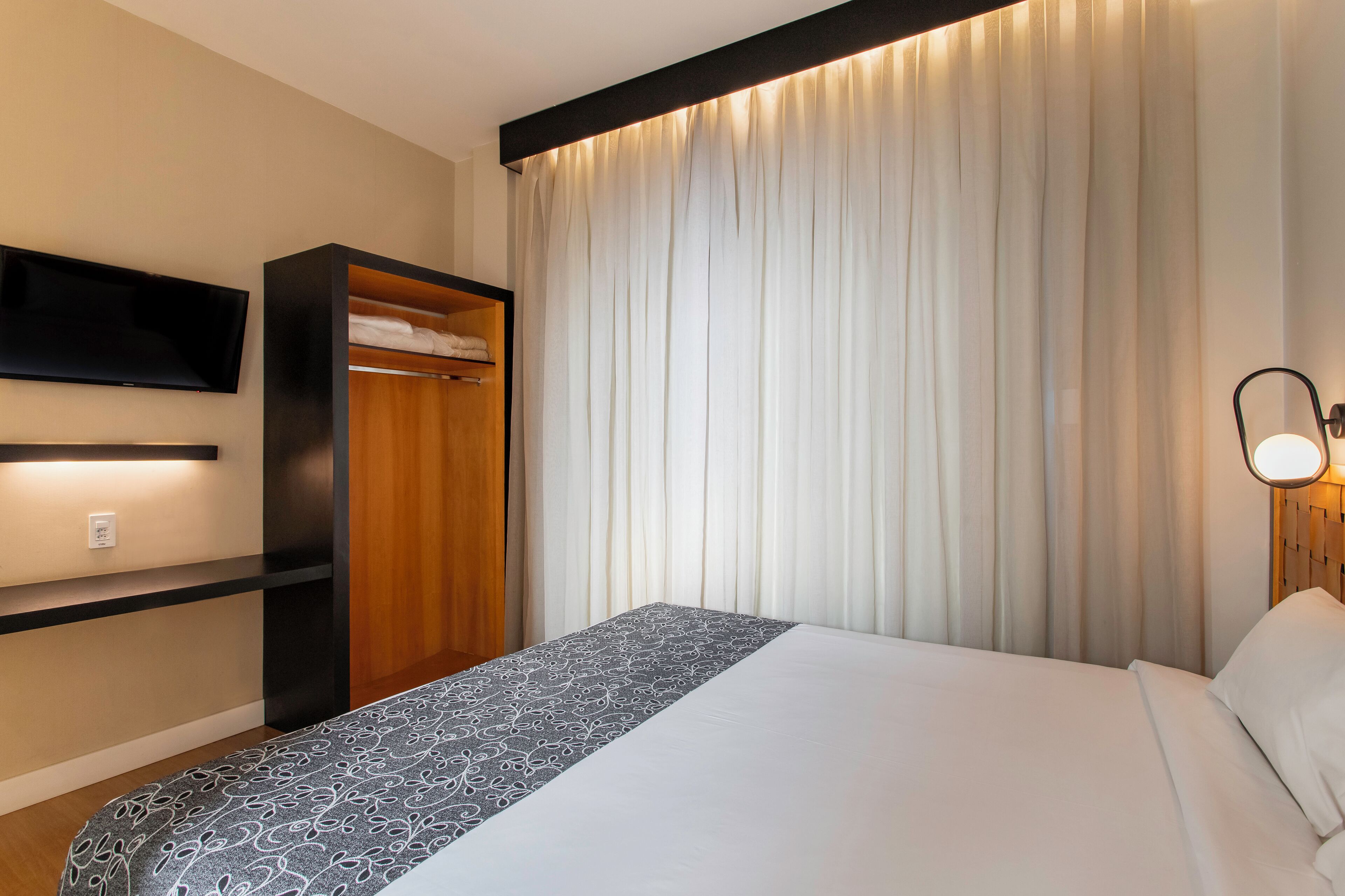 Una habitación de hotel con cama, televisor montado en la pared, armario de madera y lámpara de noche.