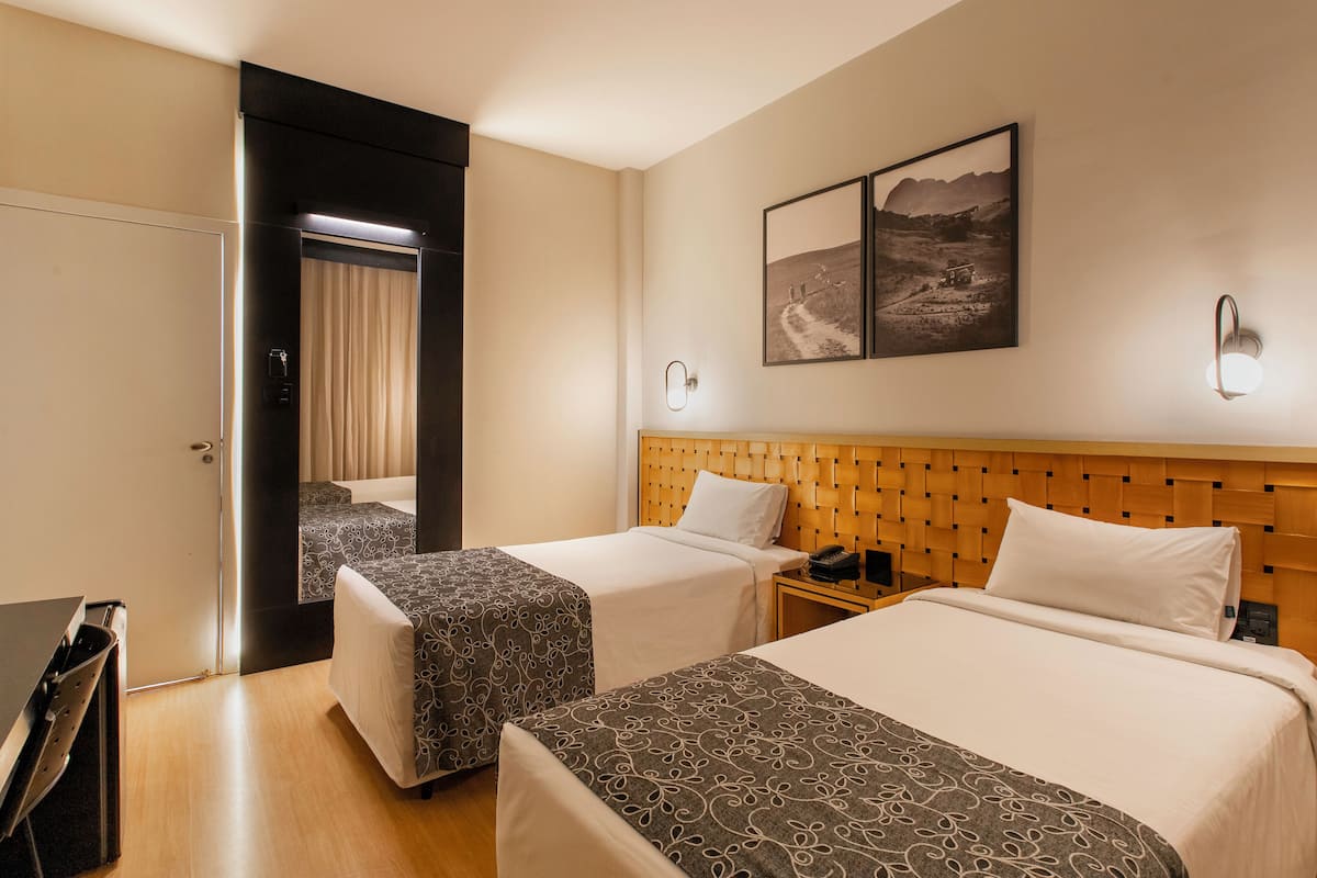 Een hotelkamer met twee bedden, een houten hoofdeinde, een nachtkastje, een lamp en een deur naar een andere kamer.